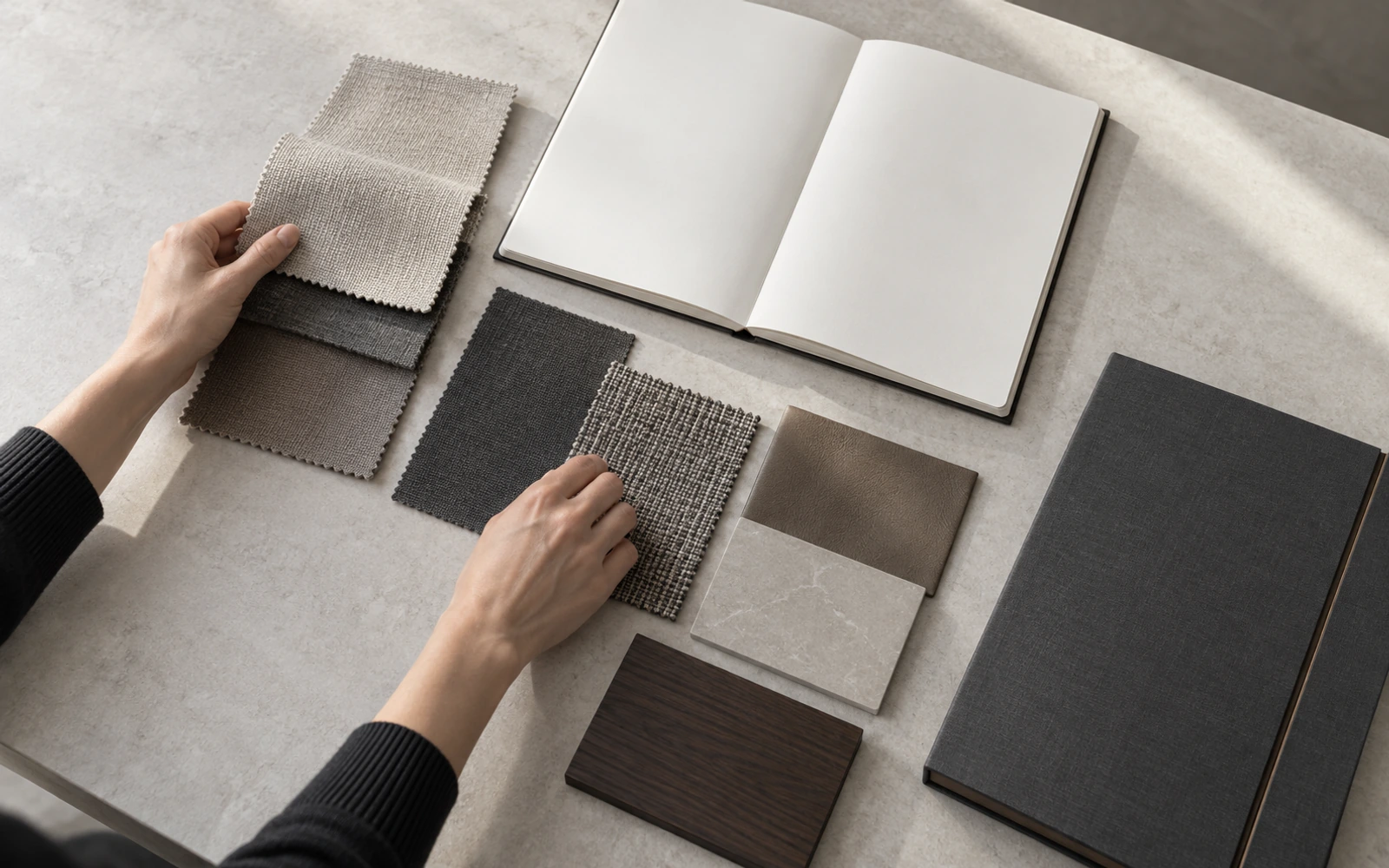 Hands arranging material swatches beside an open notebook and a sample folder on a worktable.